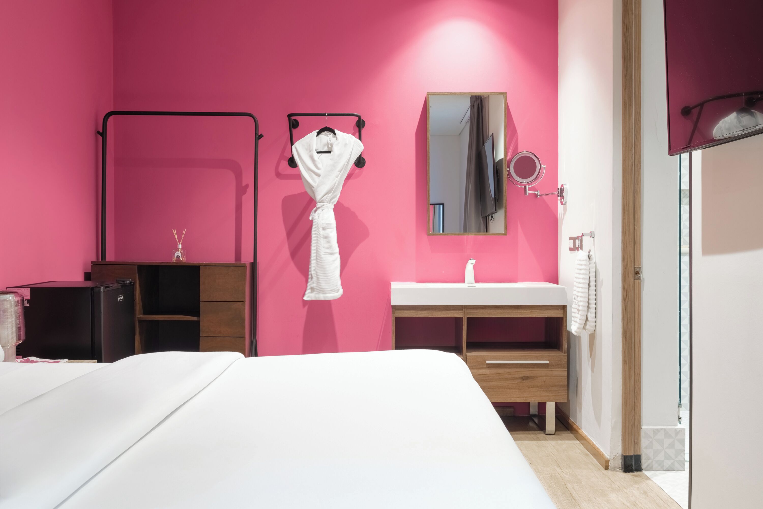 Ein Hotelzimmer mit einer rosafarbenen Wand, einem weißen Bett, einer Holzkommode, einem weißen Schminktisch und einem Spiegel.