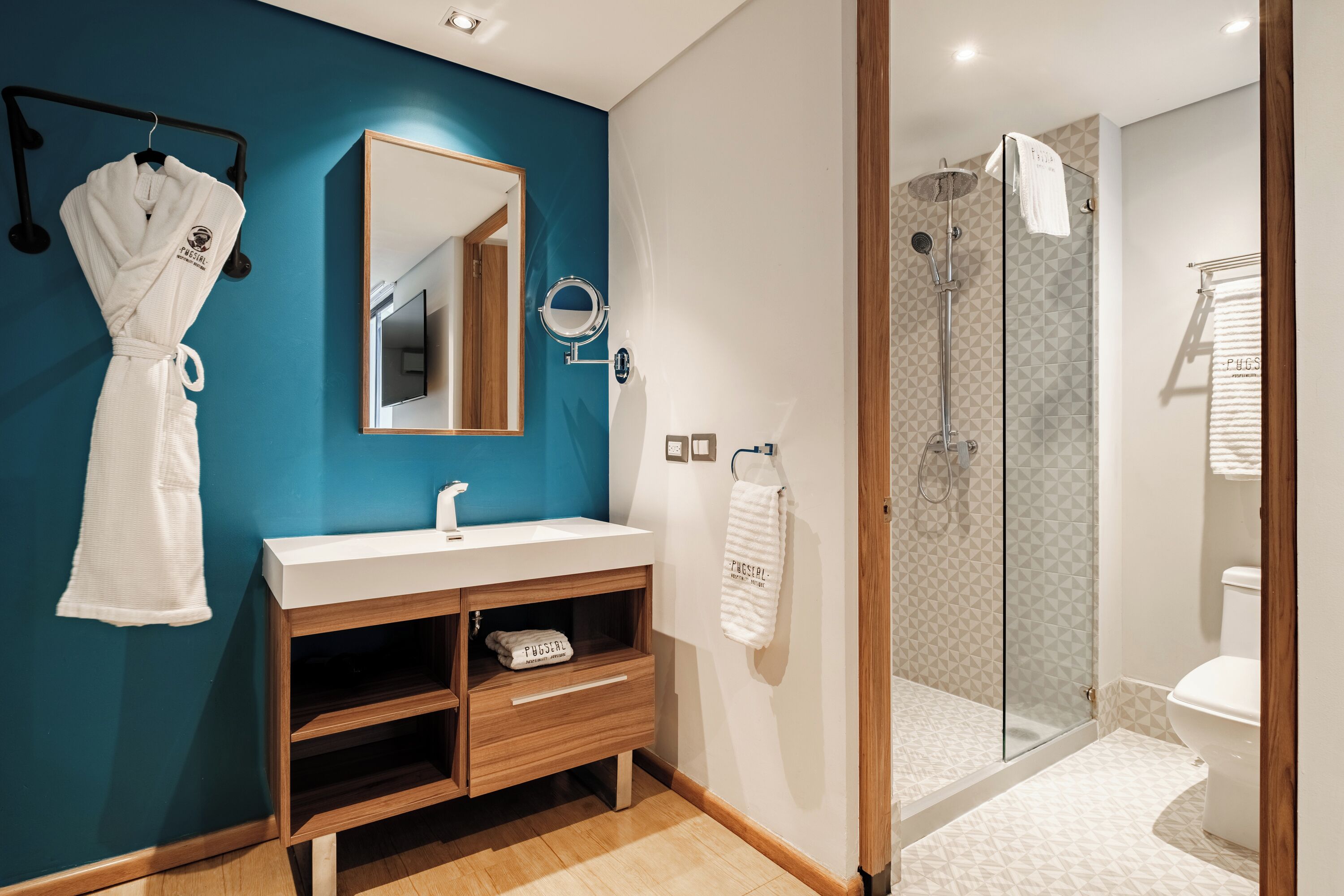 A modern bathroom with a blue accent wall, a white sink vanity, a glass shower enclosure, and a hanging robe.