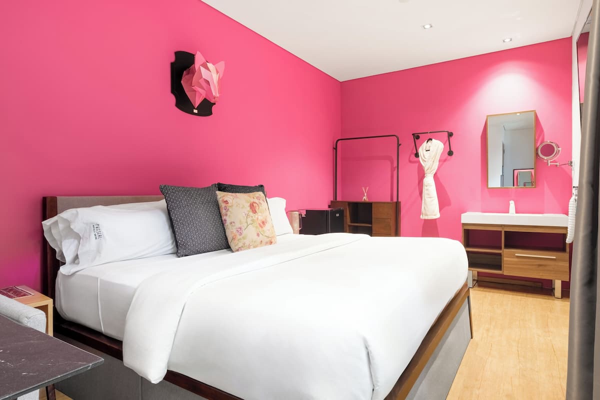 A hotel room with a large bed, a wooden nightstand, a vanity with a mirror, and a pink wall.