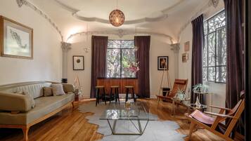 Ein Wohnzimmer mit einer Couch, einem Glastisch und Holzboden. Zwei Fenster mit Vorhängen, eine Chandelieber und ein Gemälde an der Wand sind zu sehen.