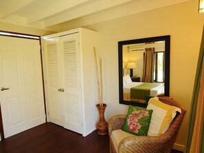A room with a closed white door, a wicker chair with a pillow, a mirror on the wall, and a tall vase with sticks.