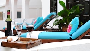 A sunlit outdoor lounge area with a wooden table holding two wine glasses and a bottle, blue cushioned lounge chairs, and a potted plant. - Villa Madeleine (Micoud)