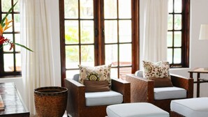 A room with wicker furniture, a wooden table, and a large window with curtains. - Villa Madeleine (Micoud)