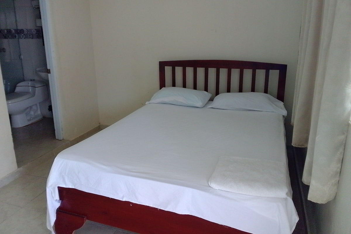 A small, single-bed room with a wooden headboard, white bedding, and a visible bathroom with a toilet and sink.