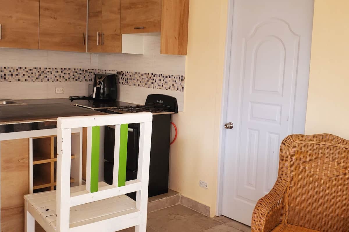 A compact kitchen with wooden cabinets, a white door, and a wicker chair.