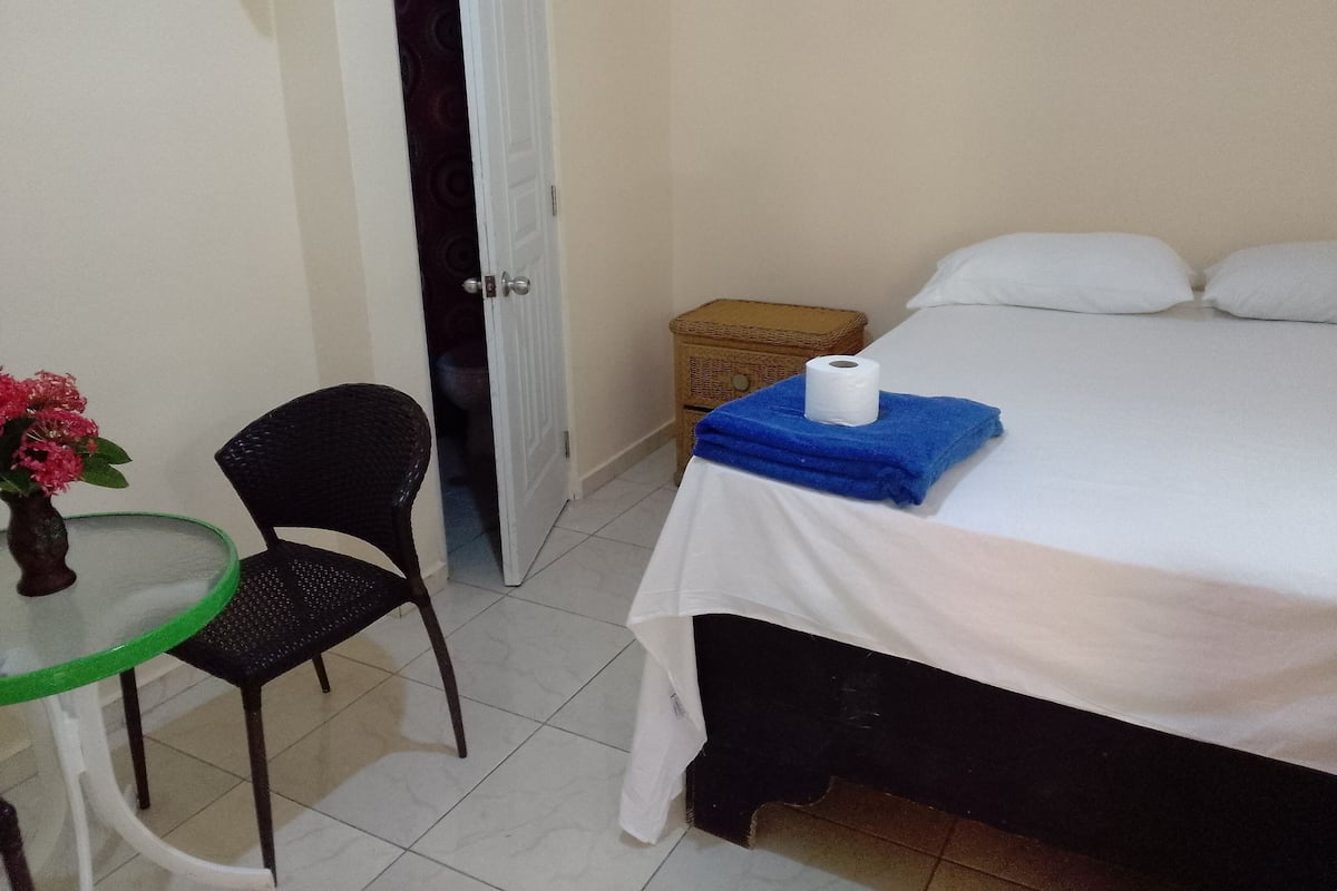 A hotel room with a bed, a small table with a chair, a vase with flowers, and a door leading to a bathroom.