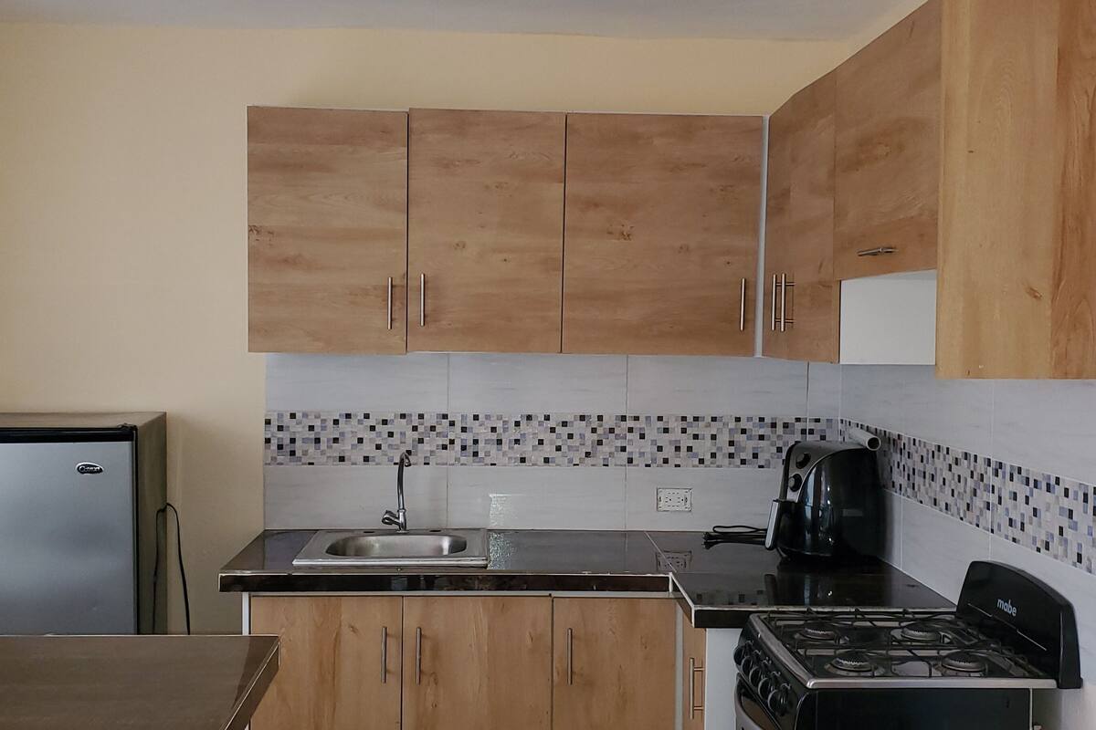 A compact kitchen with wooden cabinets, a stainless steel refrigerator, a black countertop, a sink, a stove, and a coffee maker.
