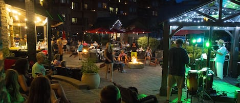 An outdoor seating area with tables and chairs, a stage with musical instruments, and a building in the background.