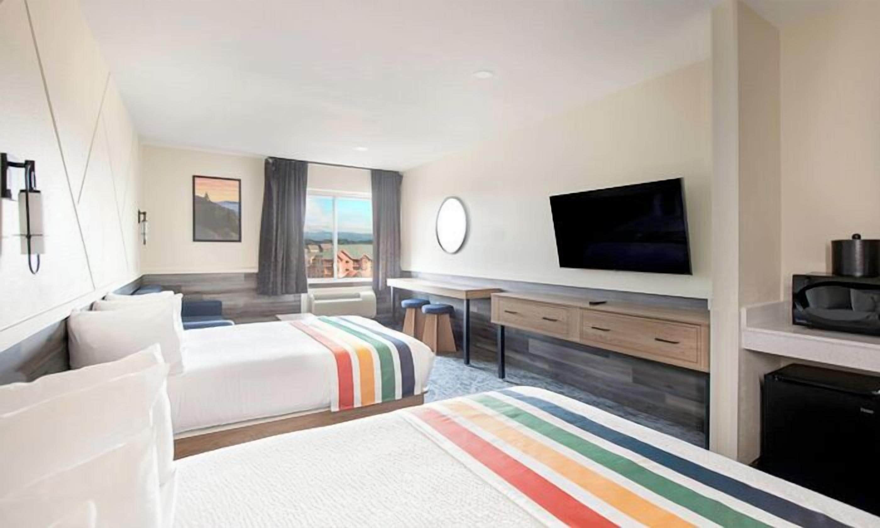 A hotel room with two beds, a desk, a television, and a view of the outside.