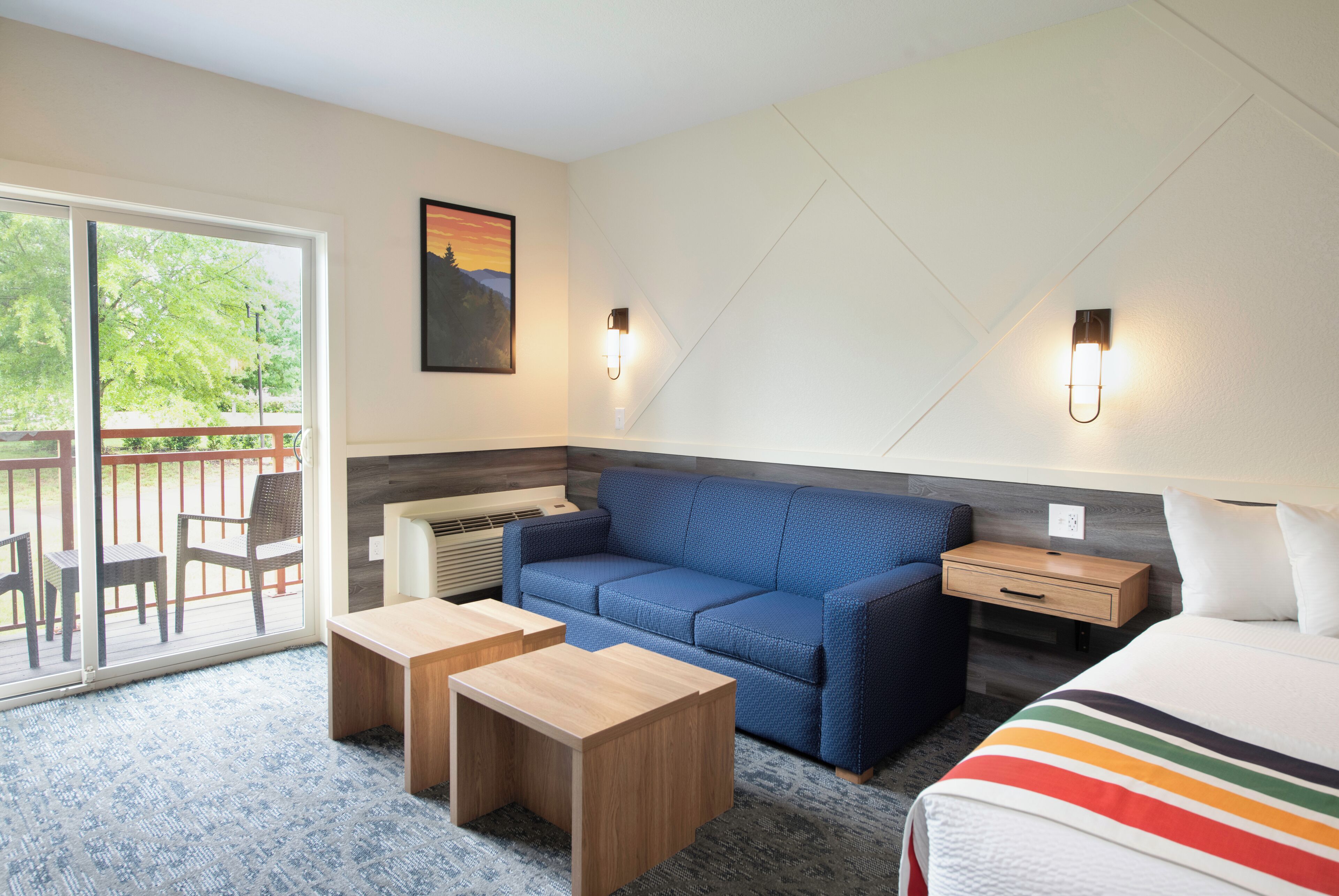 A modern hotel room with a blue sofa, a wooden nightstand, a small table, and a bed with a striped blanket.