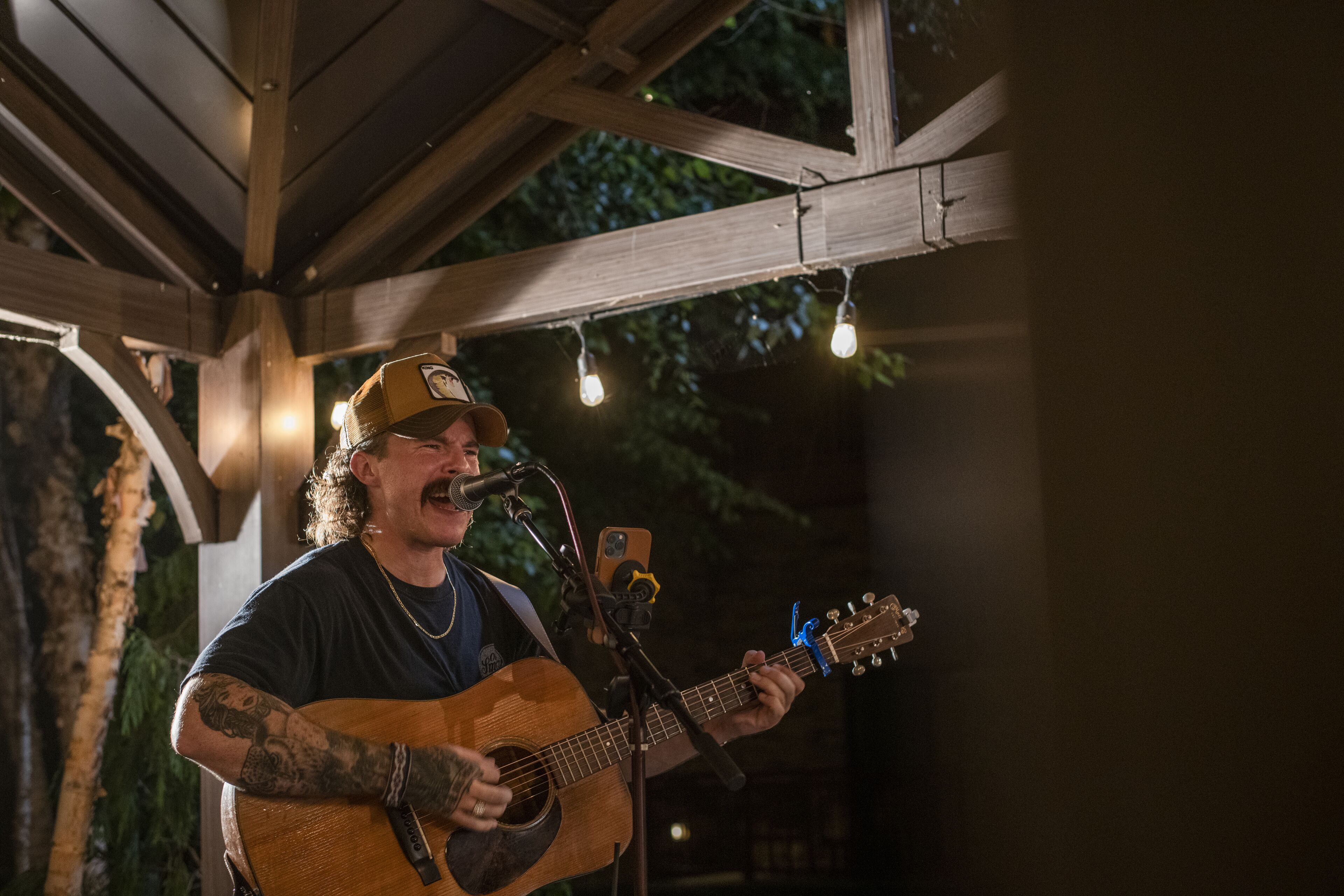 A person playing an acoustic guitar under a wooden structure with string lights.