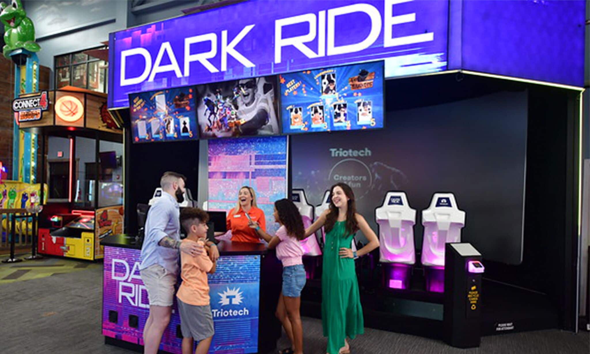 Een gezin in een speelhal genaamd 'Dark Ride' met een 'Triotech' bord, met verschillende spelautomaten en een neonreclame.