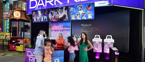 A family at a gaming arcade named 'Dark Ride' with a 'Triotech' sign, featuring various gaming machines and a neon sign.