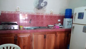 A kitchen with a gas stove, sink, wooden cabinets, a fan, a refrigerator, and a blue water dispenser. - Hostal Miconia (Puerto Ayora)