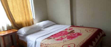 Una cama individual con sábanas blancas y una colcha de estampado floral en una habitación con una mesita de noche de madera y una ventana con cortinas.