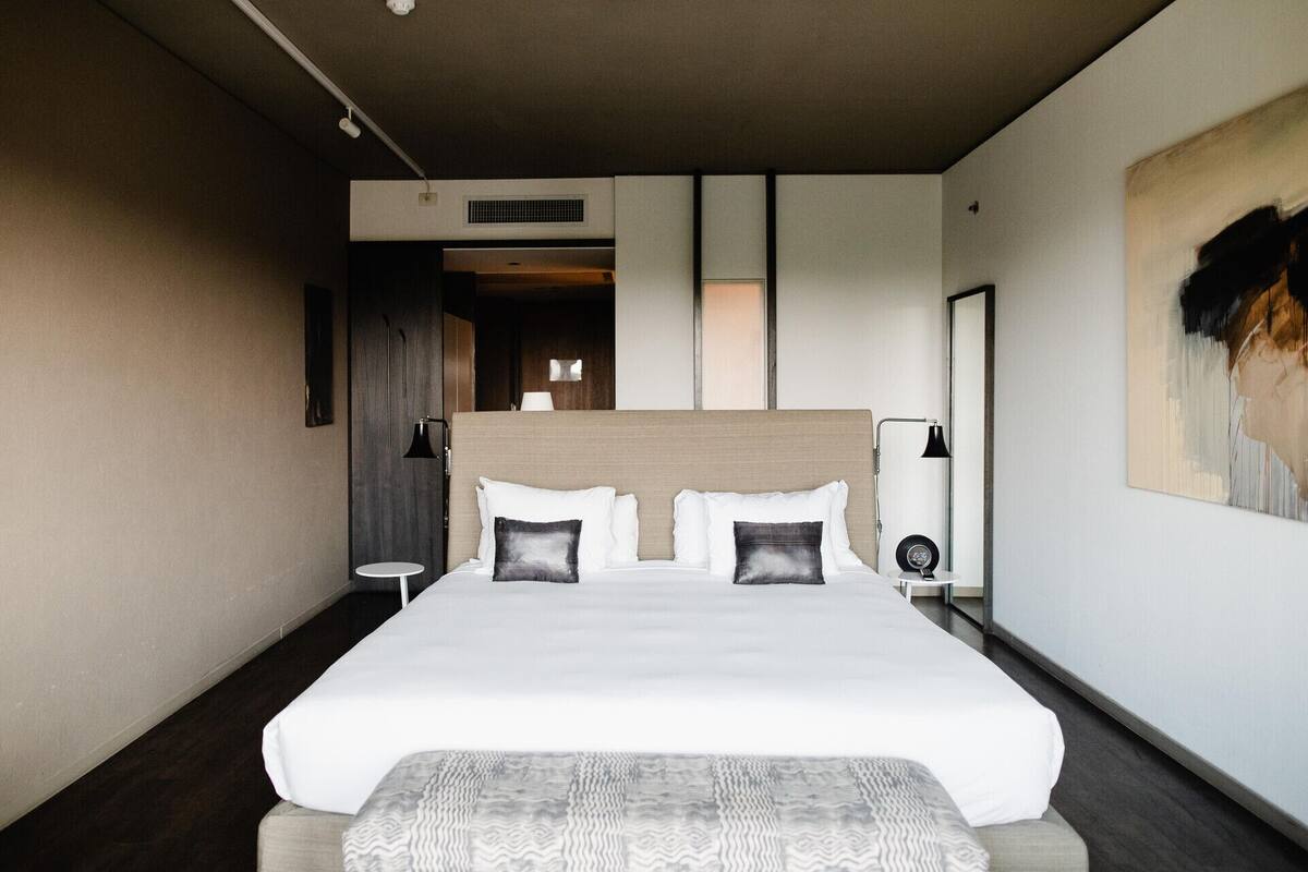 A modern hotel room with a large bed, bedside lamps, a small table, and a painting on the wall.