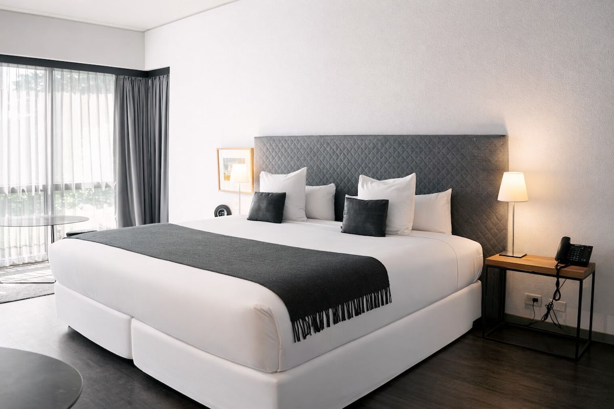 A modern hotel room with a large bed, a bedside table with a lamp, and a view of the outdoors through the window.