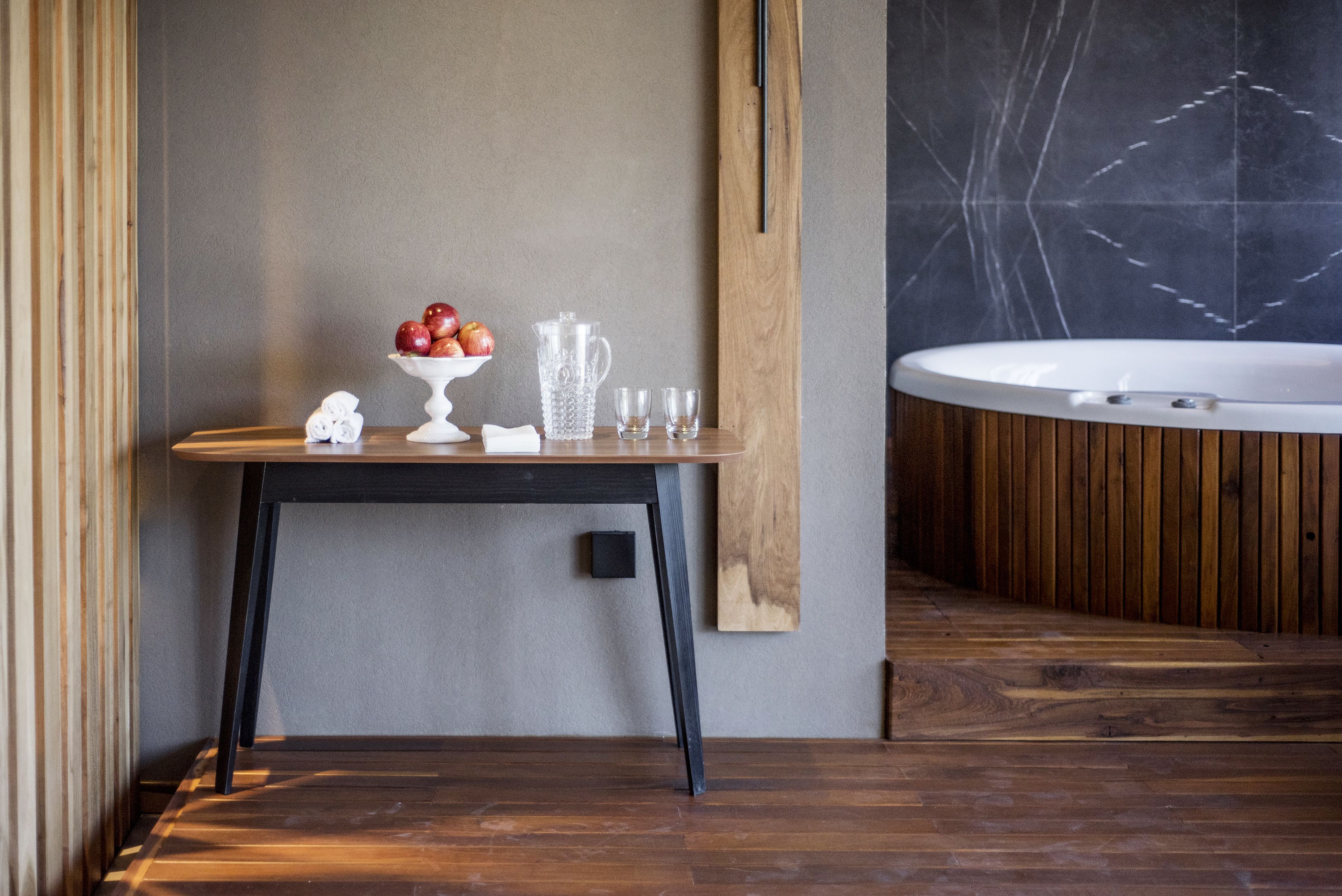 Una mesa de madera con un frutero, una jarra y vasos en un baño moderno con suelo de madera y pared de mármol.