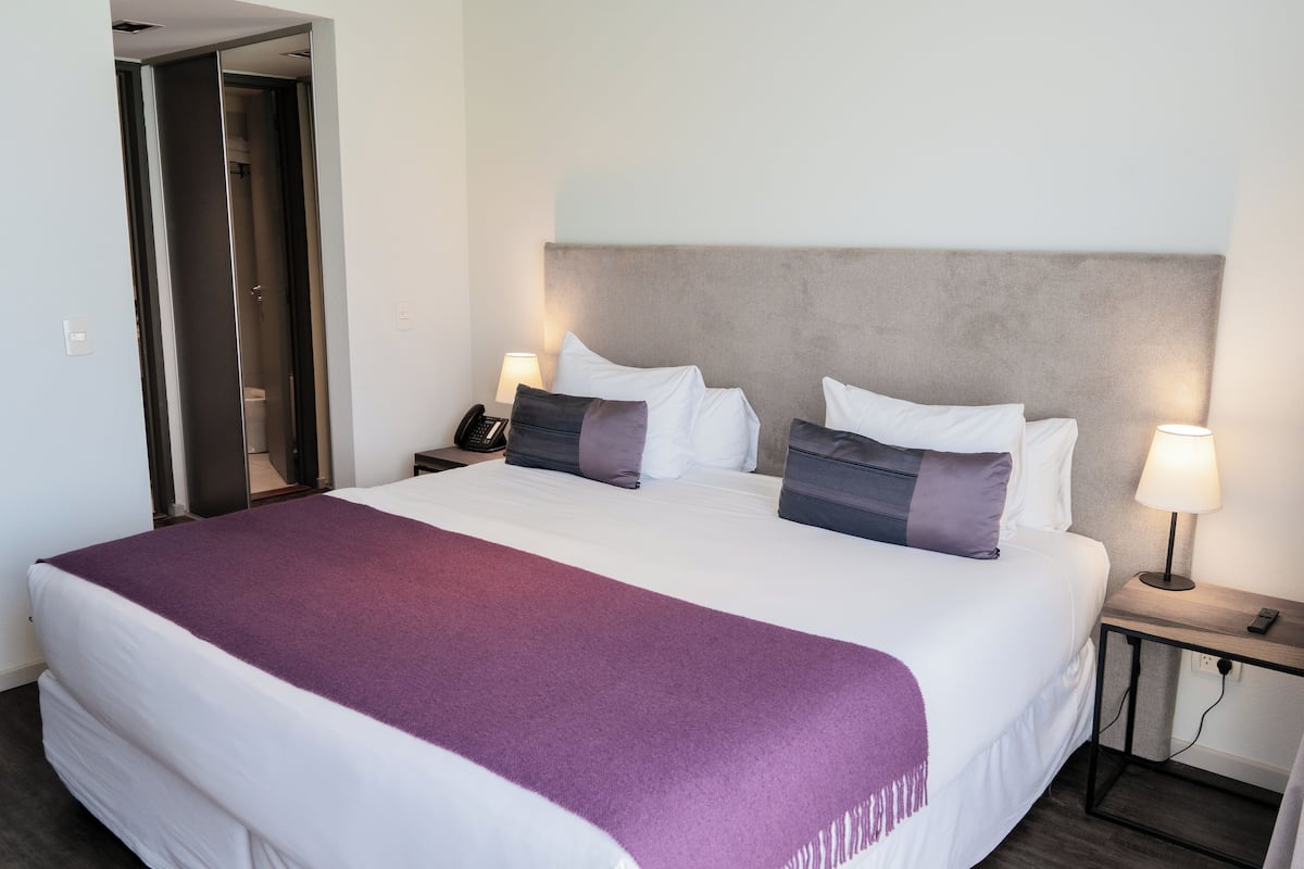 A hotel room with a large bed, a bedside table with a lamp, and a door leading to a bathroom.
