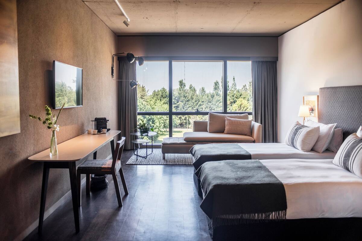 A modern hotel room with a large window, a sofa, a bed, a desk, and a TV.