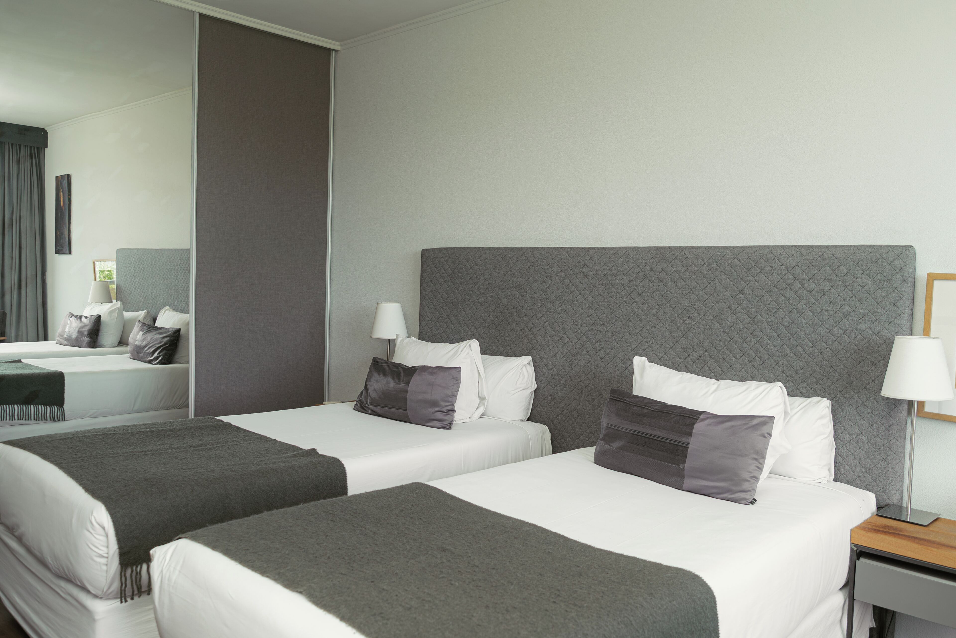 A hotel room with two beds, a large mirror, a bedside table with a lamp, and a view of the outside.
