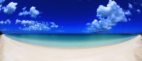 Ein Strand mit klarem türkisblauem Wasser und einem weiten, sandigen Ufer unter einem strahlend blauen Himmel mit vereinzelten Wolken.