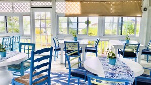 A covered outdoor seating area with blue chairs and white tables, featuring potted plants and a view of greenery. - Rollezz Villas Beach Resort (Old Bight)