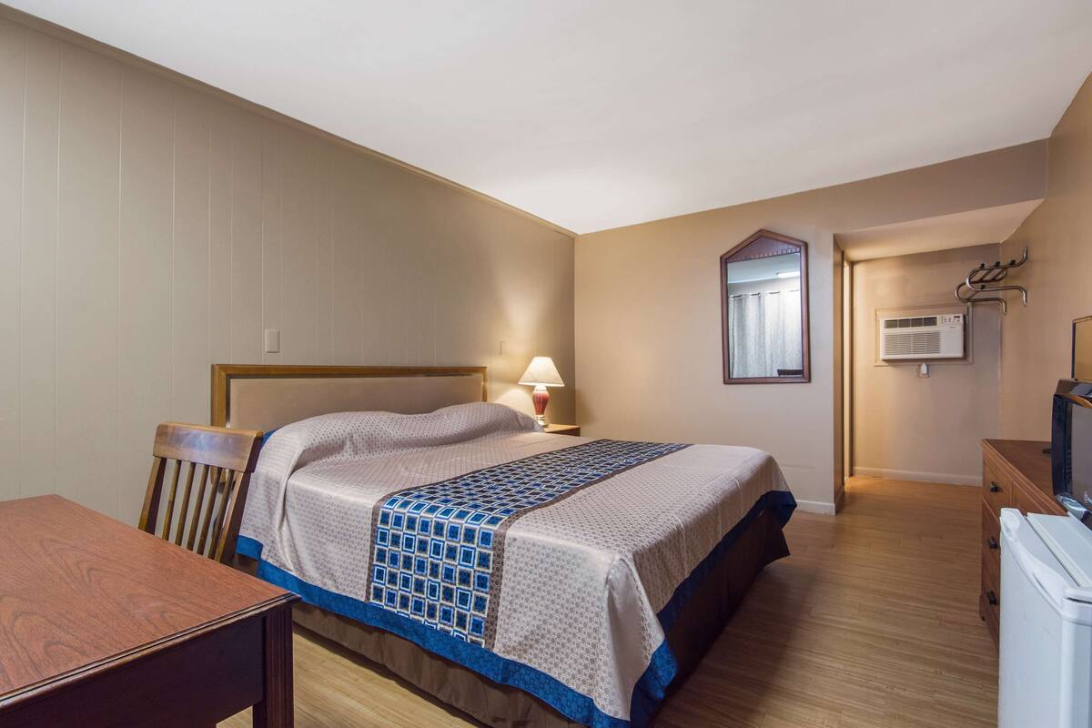Quarto com cama, cadeira de madeira, mesa, luminária, cômoda e aparelho de ar condicionado.