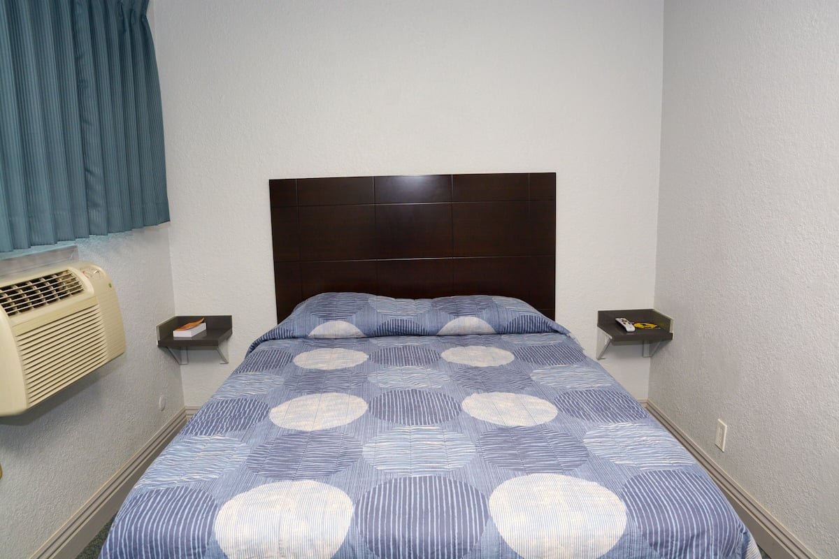 A hotel room with a bed, a nightstand, a remote control, and a wall-mounted air conditioner.