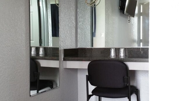Habitación de hotel con un espejo grande, una silla negra, un televisor de pantalla plana montado en la pared y una rejilla de ventilación en el techo.
