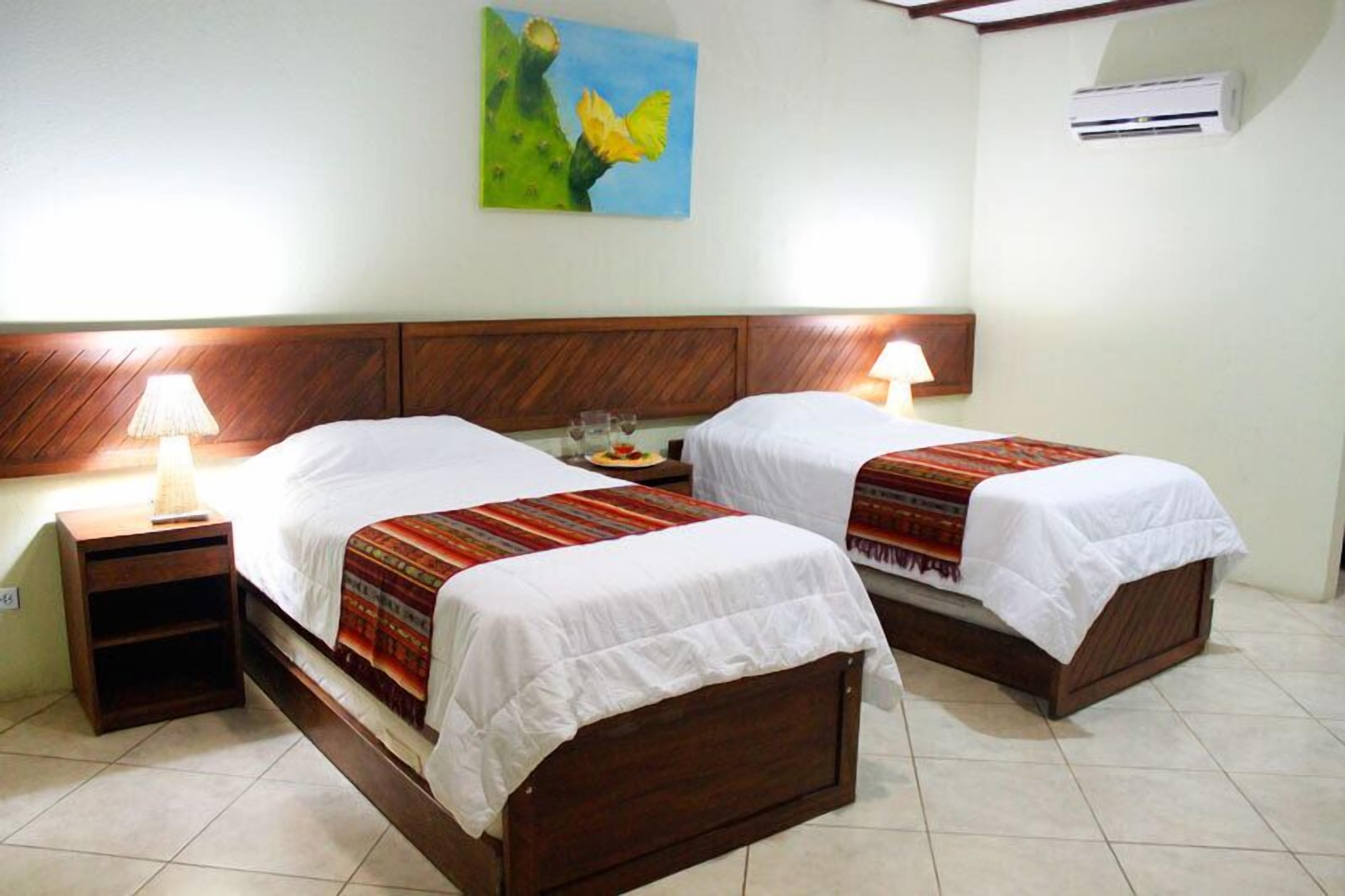 A hotel room with two wooden beds, each with a white bedspread and a colorful blanket. There is a nightstand with a lamp on the left side, and an air conditioning unit on the wall.