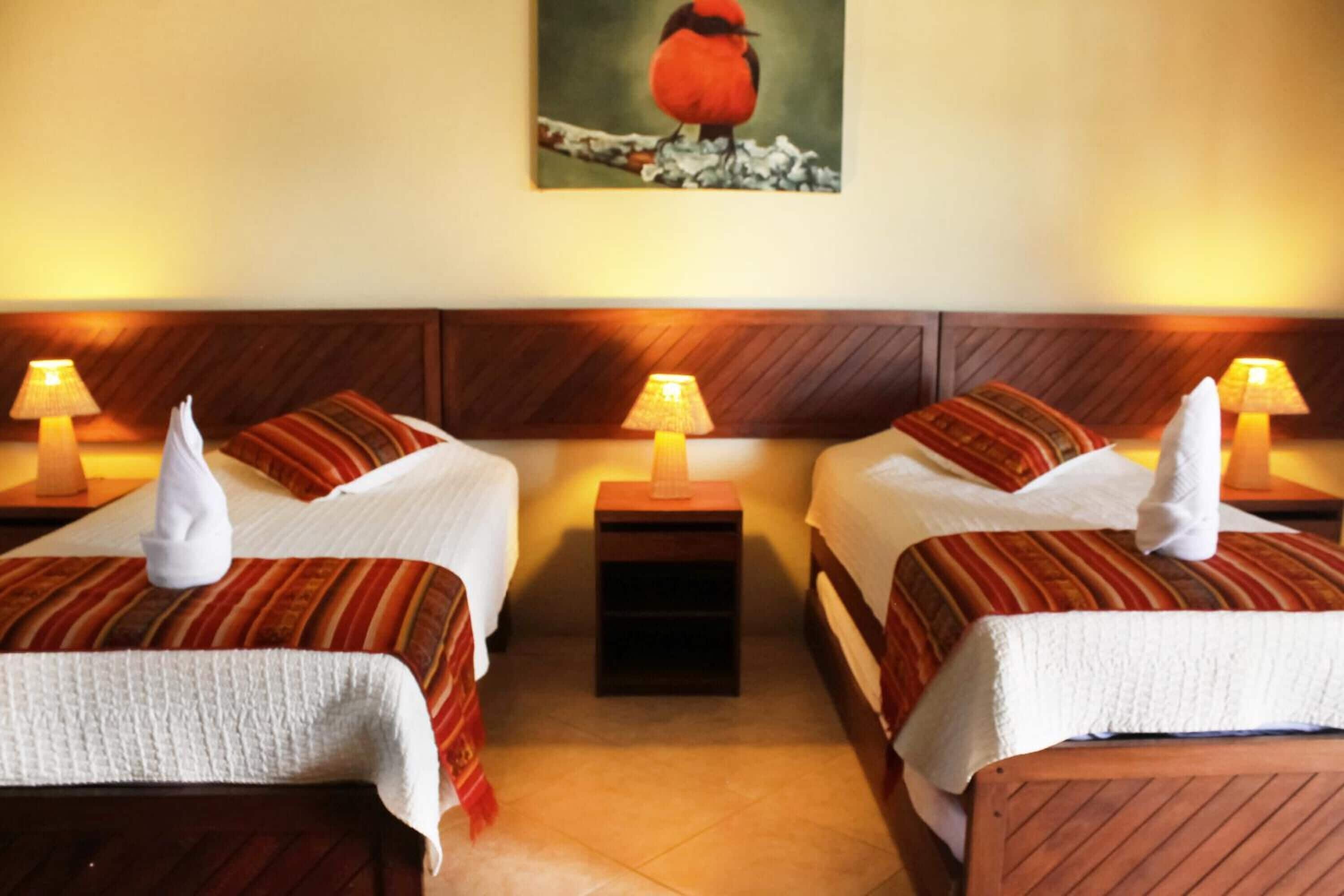 Habitación de hotel con dos camas, cada una con un almohadón y una lámpara. En la pared, un cuadro de un pájaro.