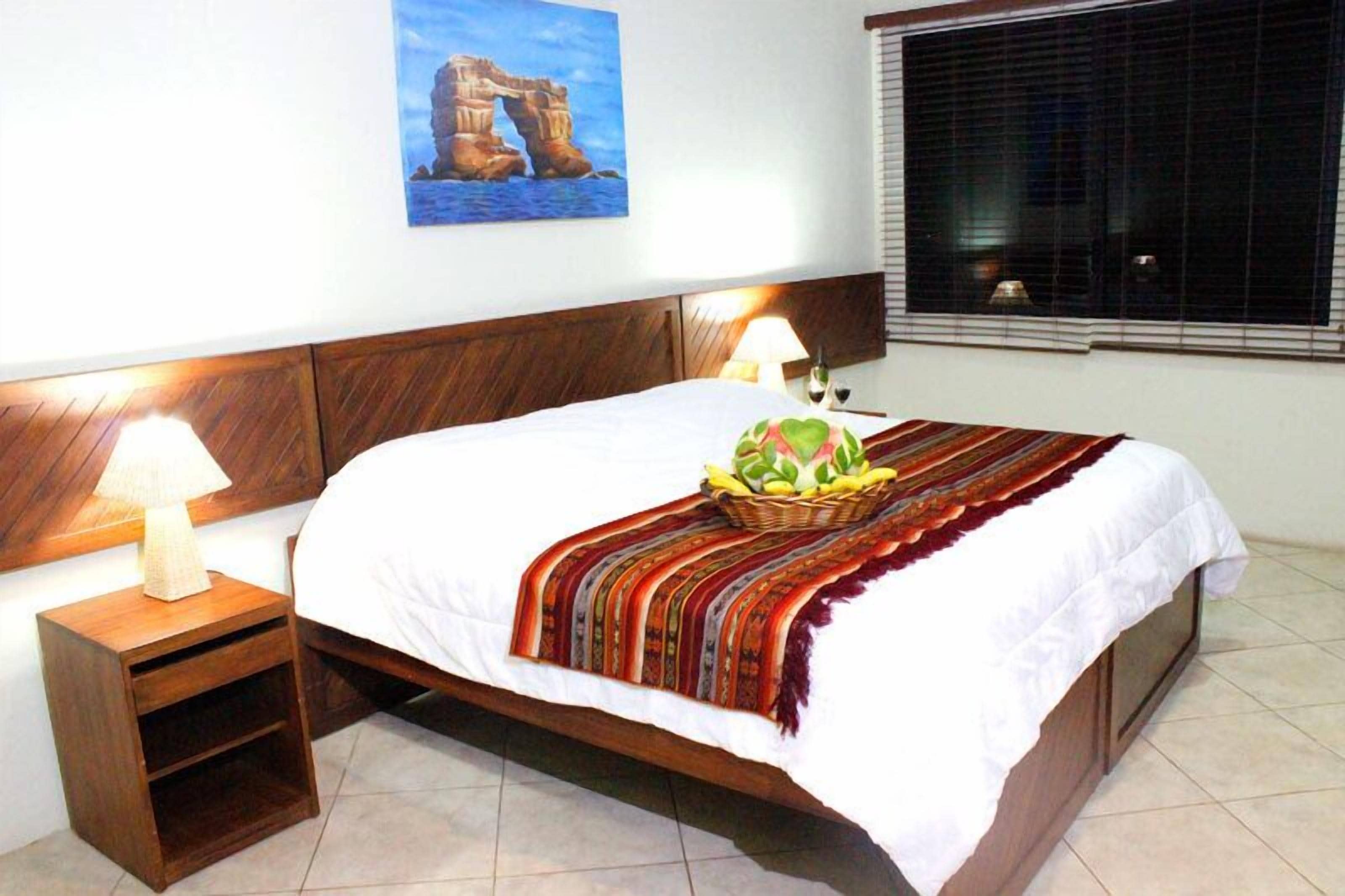 Un lit bien fait, avec une couette blanche et une couverture tissée aux couleurs vives, un panier de fruits posé sur le lit, et une lampe sur une table de chevet en bois à côté.