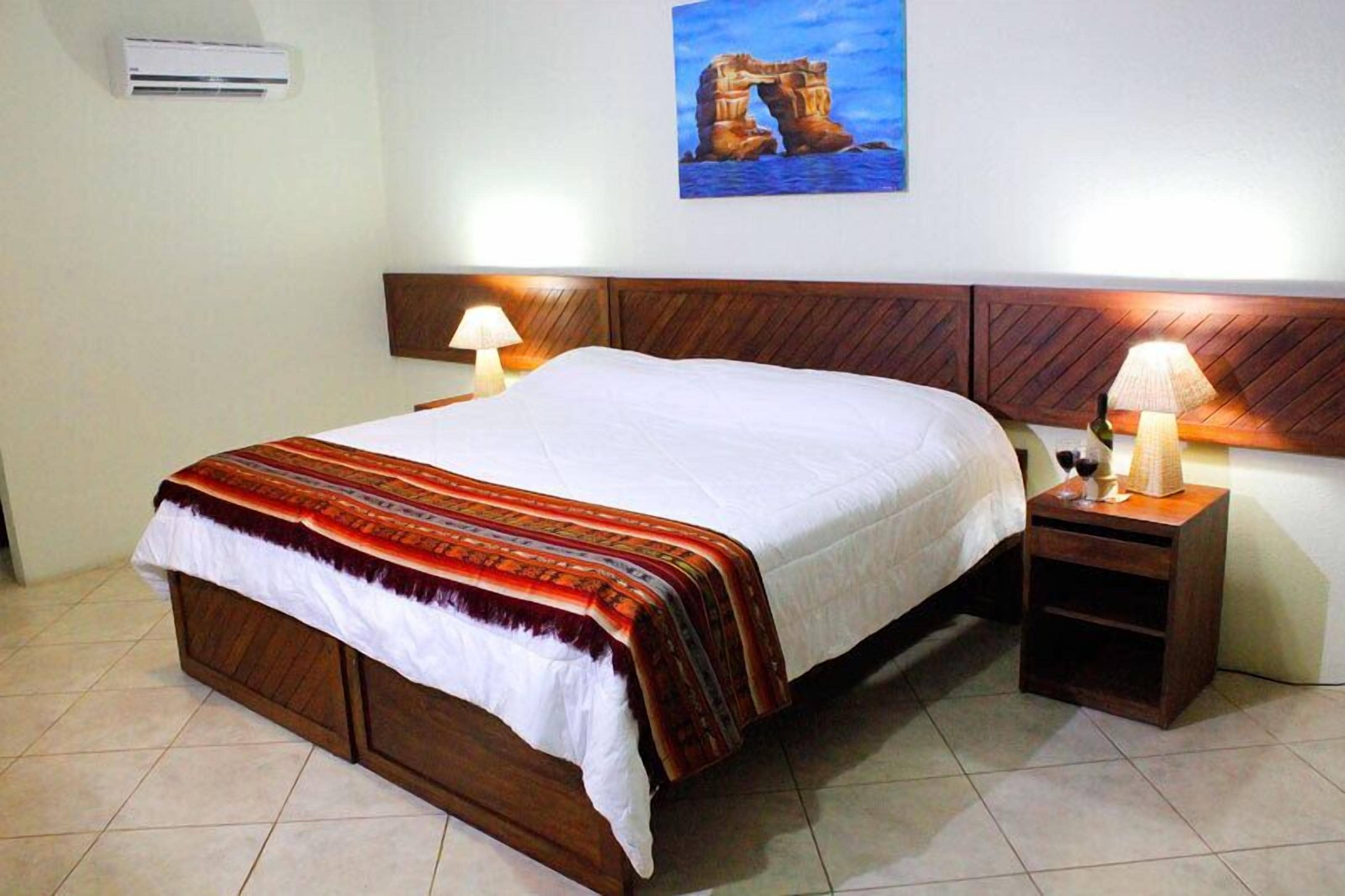 Una cama bien hecha con cabecera de madera, dos lámparas de noche y un aire acondicionado de pared.