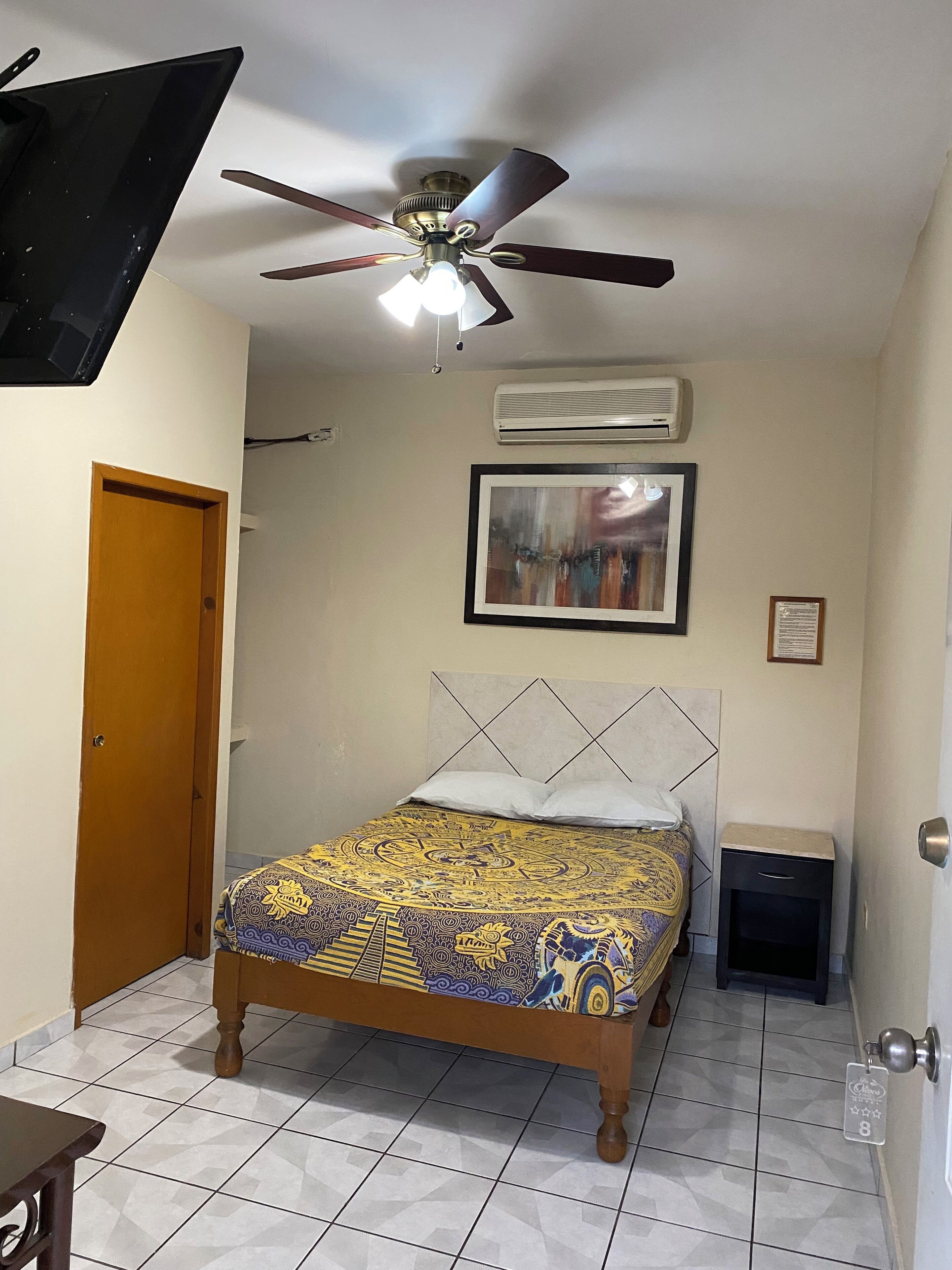A bedroom with a bed, a ceiling fan, an air conditioner, a framed picture, a door, and a small table.