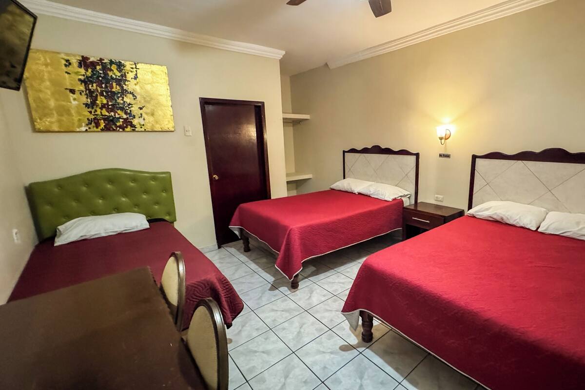 A hotel room with three single beds, a green headboard, a wooden nightstand, and a wall-mounted light fixture.