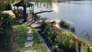 A wooden deck with outdoor furniture overlooking a lake, surrounded by lush greenery and a stone pathway. - Cottage Lake Bed and Breakfast (Woodinville)