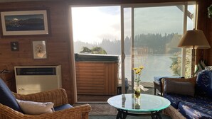 A cozy living room with wicker furniture, a glass table with flowers, and a view of a lake through a sliding door. - Cottage Lake Bed and Breakfast (Woodinville)