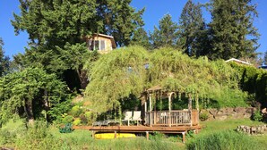 A wooden deck with chairs, a small gazebo, and a red canoe on a calm body of water, surrounded by lush greenery and trees. - Cottage Lake Bed and Breakfast (Woodinville)