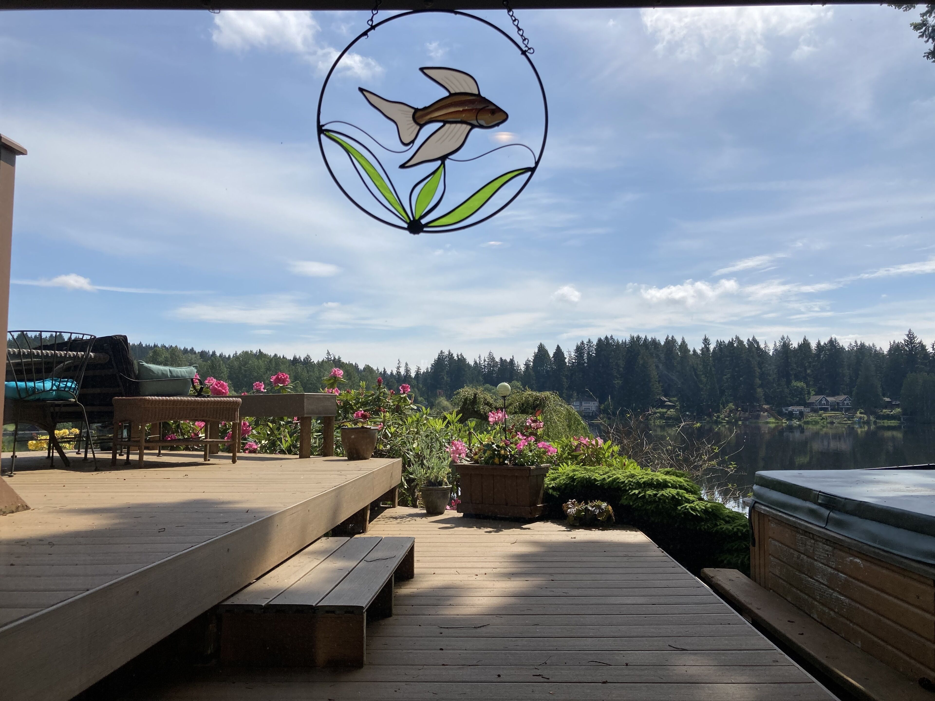 A wooden deck with benches, a hanging stained glass fish decoration, and a view of a lake and forested area.