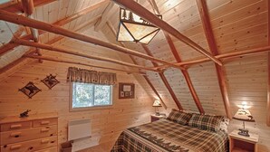 A cozy attic bedroom with a sloped ceiling, wooden beams, a bed with plaid bedding, two bedside tables with lamps, and a window with a view of trees. - Grandma's Cabin (Island Park)