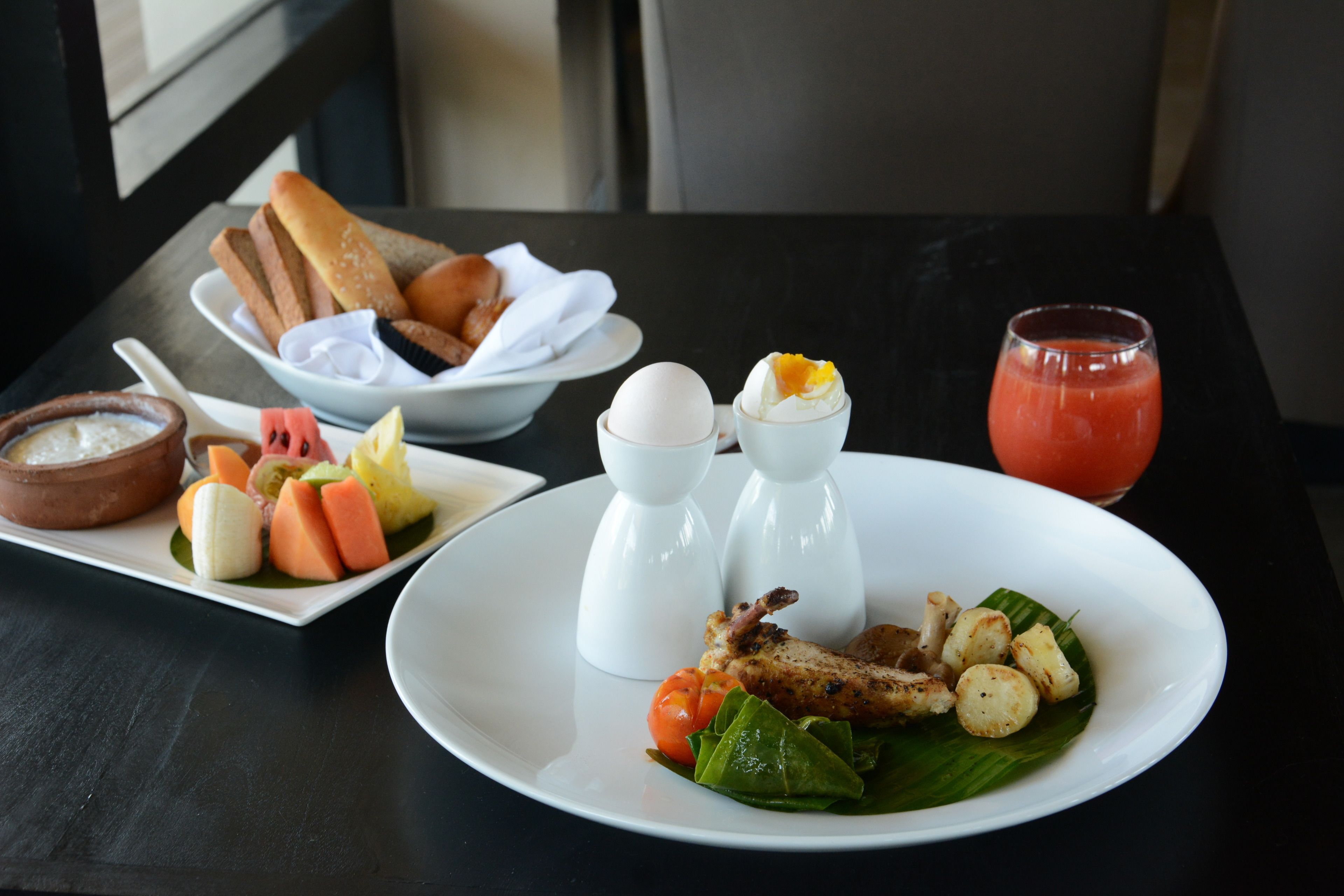 A table with a plate of food including grilled chicken, vegetables, and a fruit dessert. There is also a bowl of bread and a glass of juice.