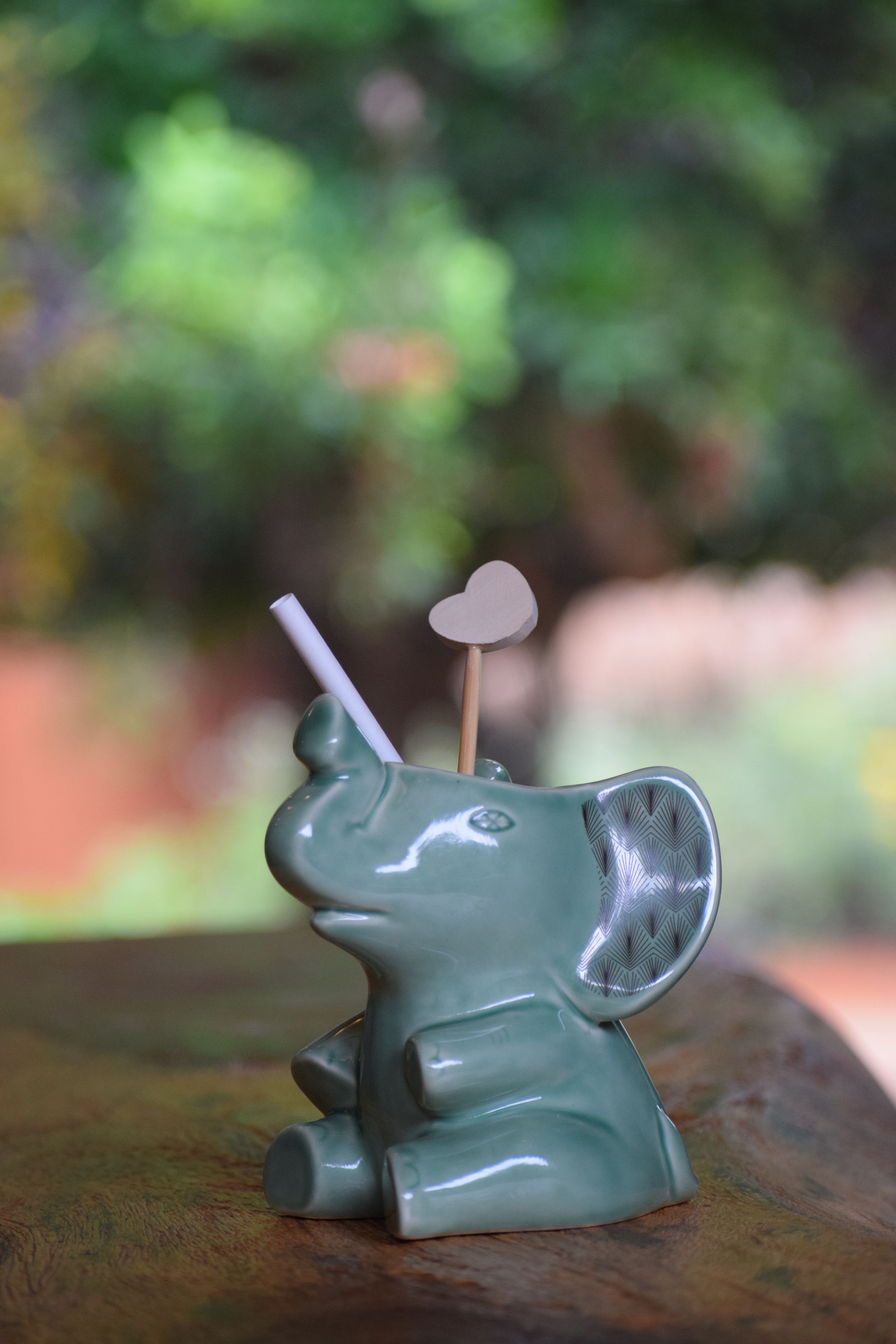 A ceramic elephant figurine with a straw and a heart-shaped object on its head, placed on a wooden surface.