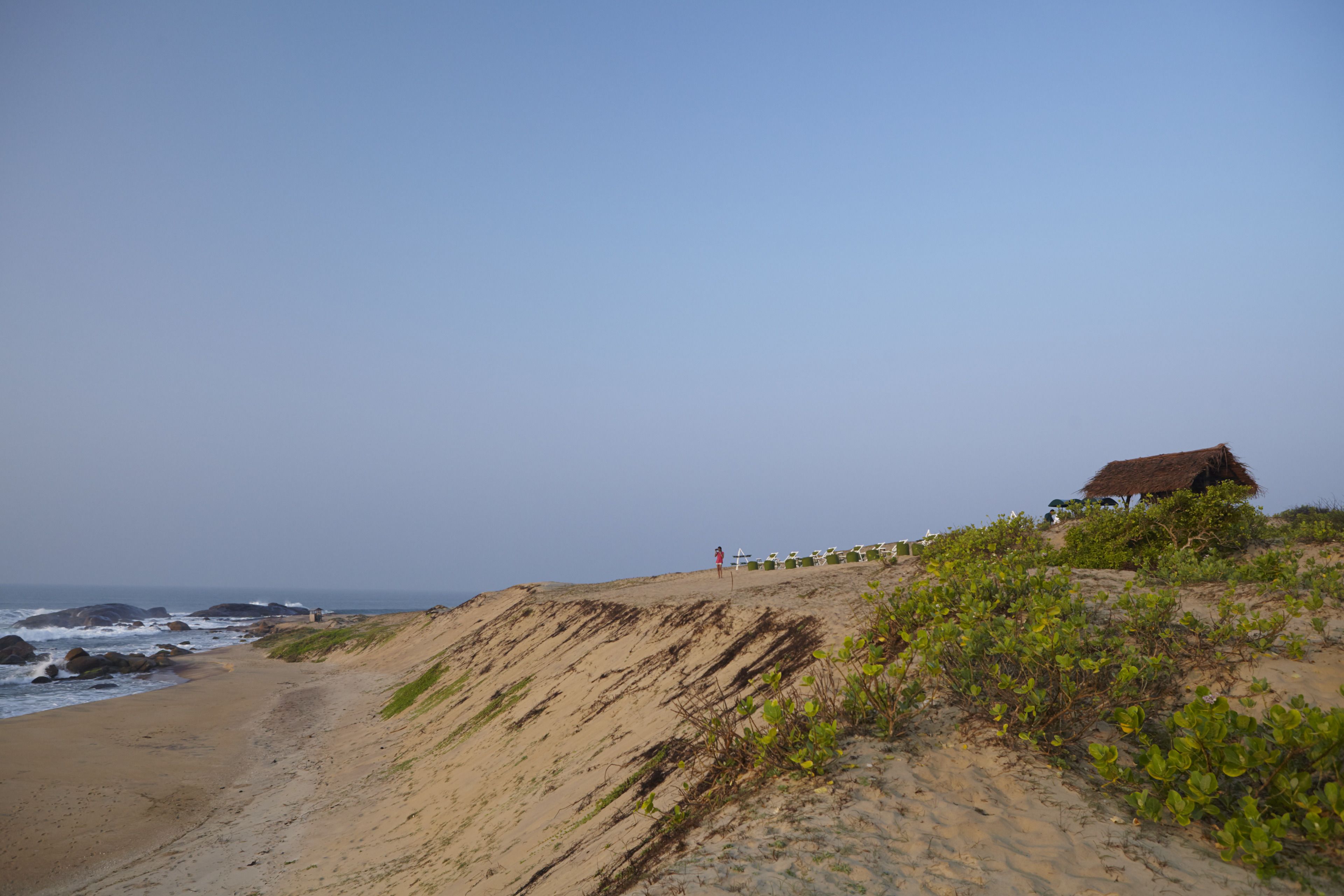 A sandy beach with a thatched-roof hut on a grassy hill, overlooking the ocean.