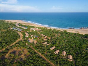 An aerial view of a coastal resort with multiple bungalow-style buildings surrounded by dense greenery, a sandy beach, and the ocean in the background. - Jetwing Jungle Lodge, A Luxury Reserve (Thissamaharama)