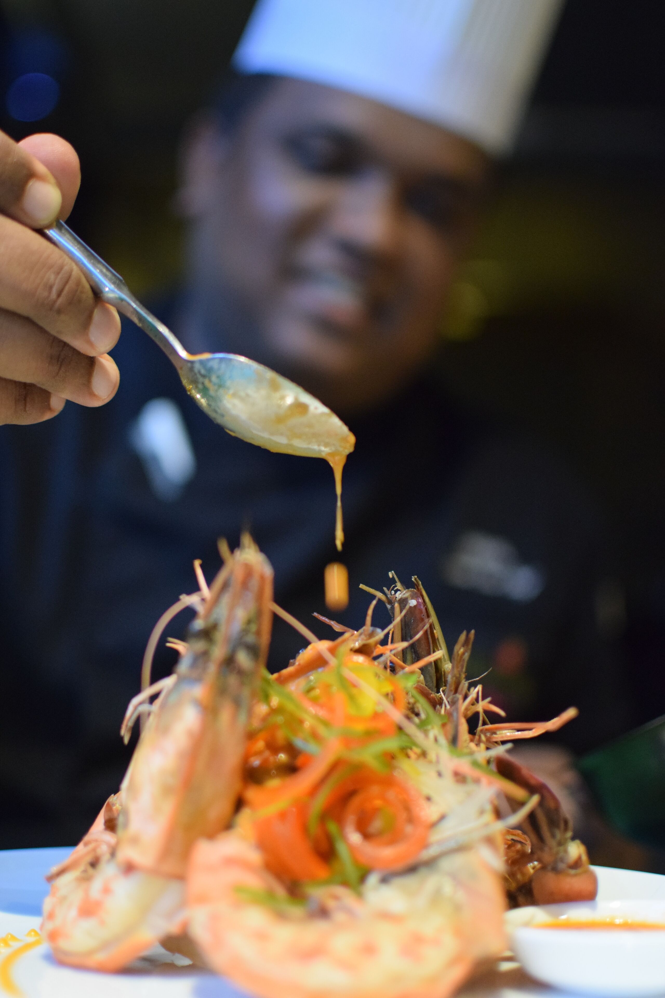 A chef holding a spoon over a dish of seafood, including shrimp and possibly lobster, with a blurred background.