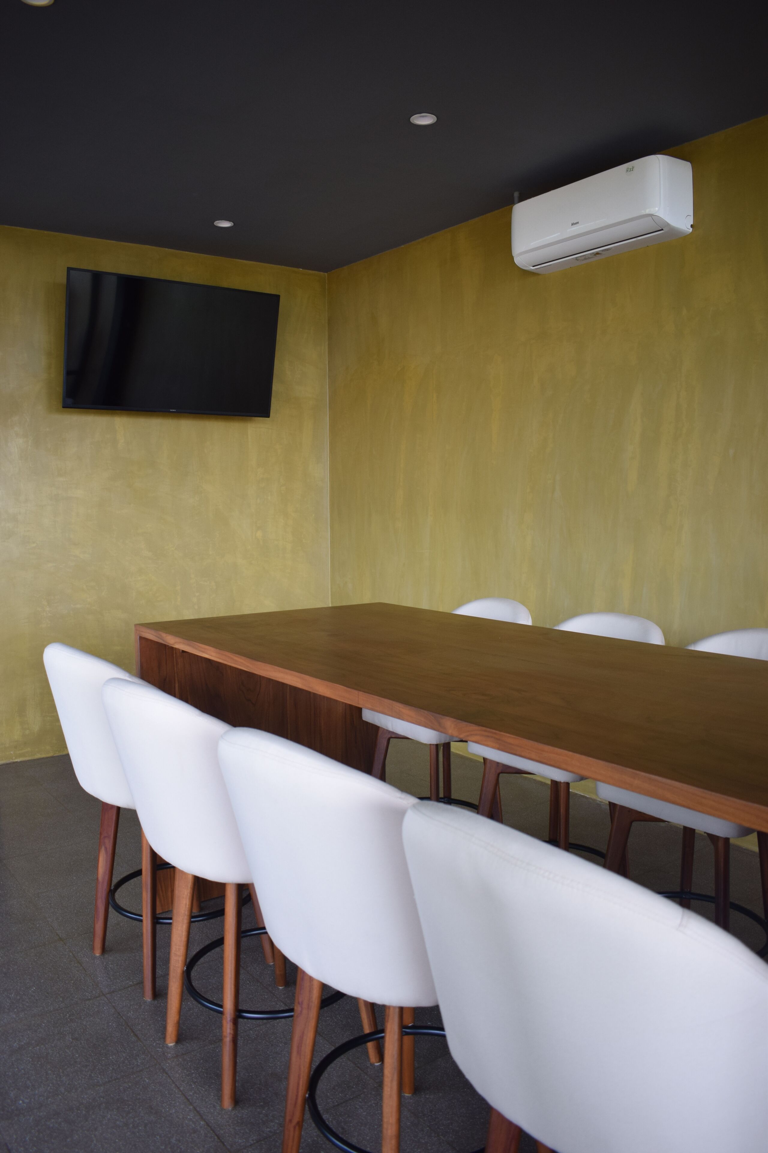 A conference room with a wooden table, white chairs, a flat-screen TV mounted on the wall, and an air conditioning unit.
