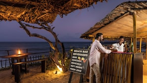 A beach bar with a sign listing beverages, a wooden bench, and a thatched roof.