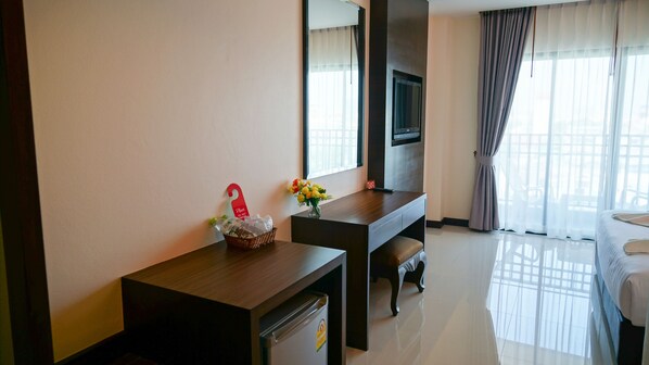 Habitación de hotel con un escritorio de madera, un televisor montado en la pared, una cama con edredón blanco y una ventana con cortinas traslúcidas.