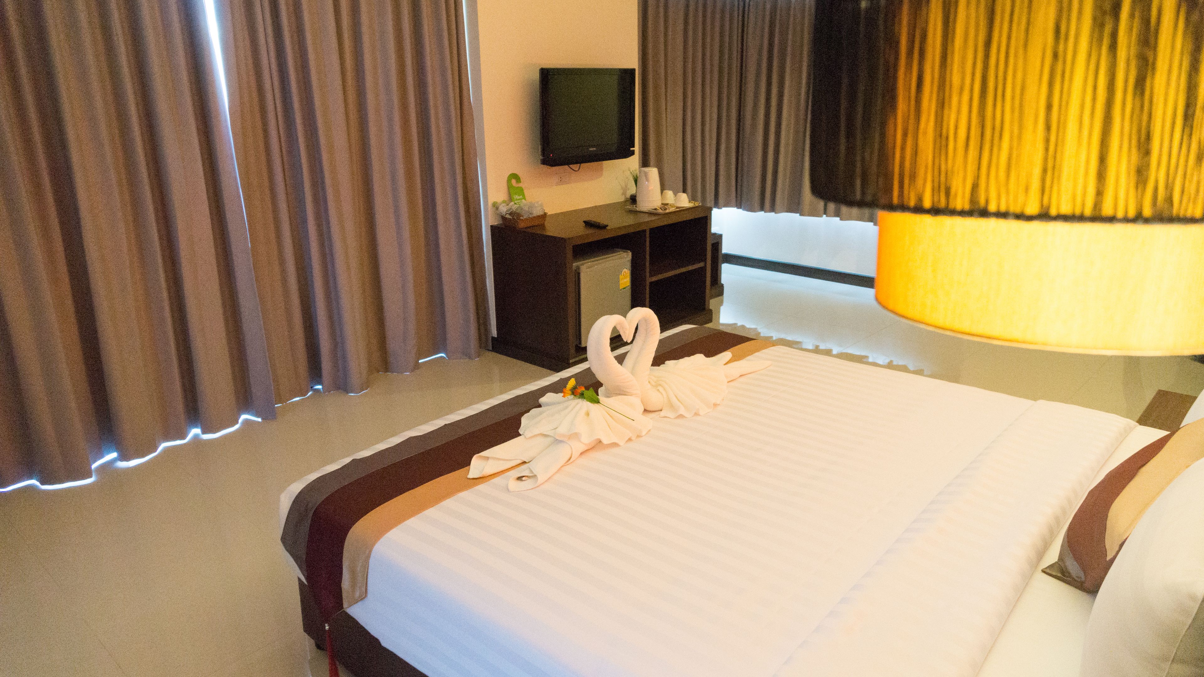 A hotel room with a neatly made bed, a television on a stand, a desk with a lamp, and curtains.